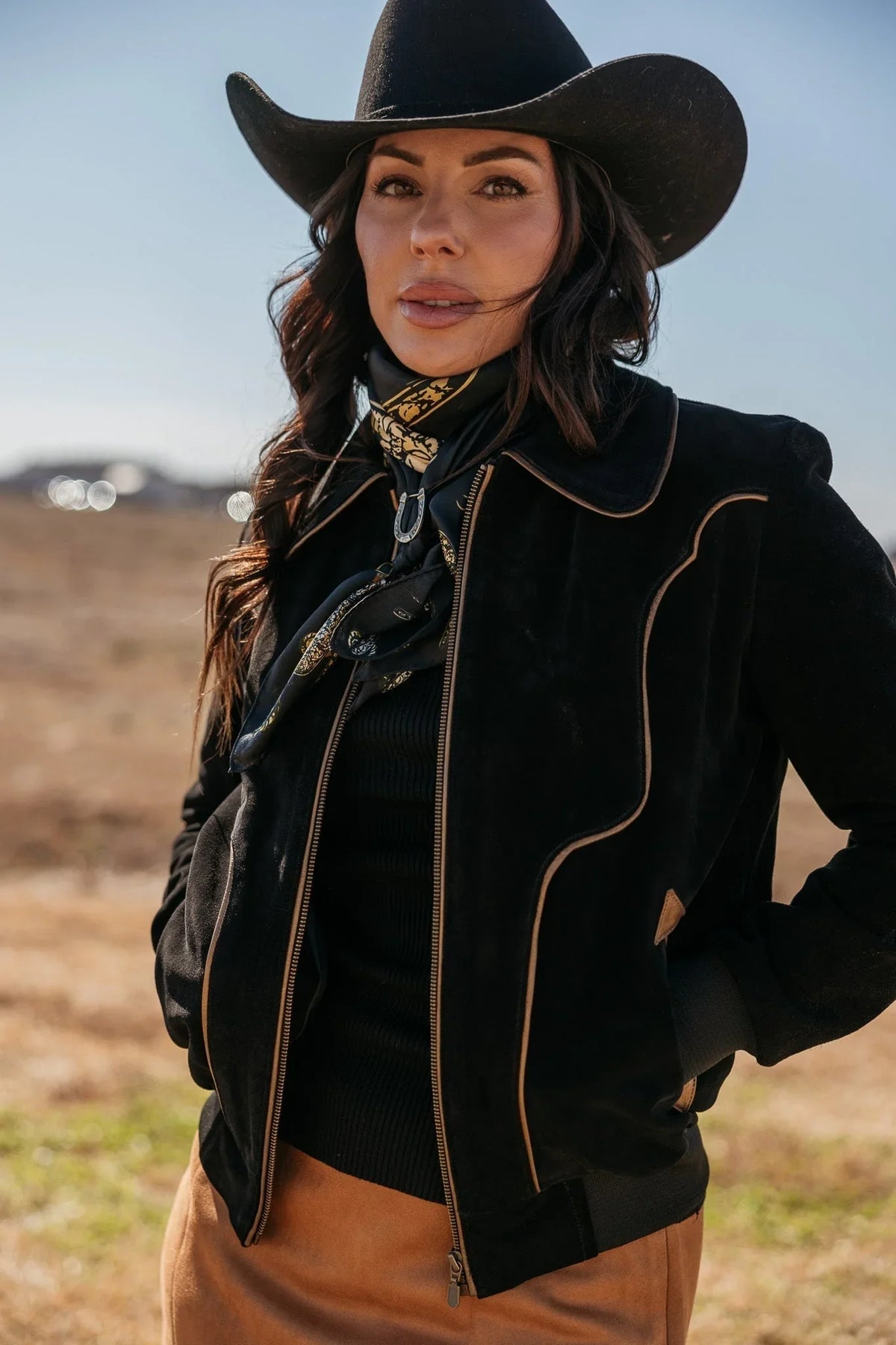 Woman in black cowboy hat, western suede jacket, and printed scarf in an outdoor setting.
