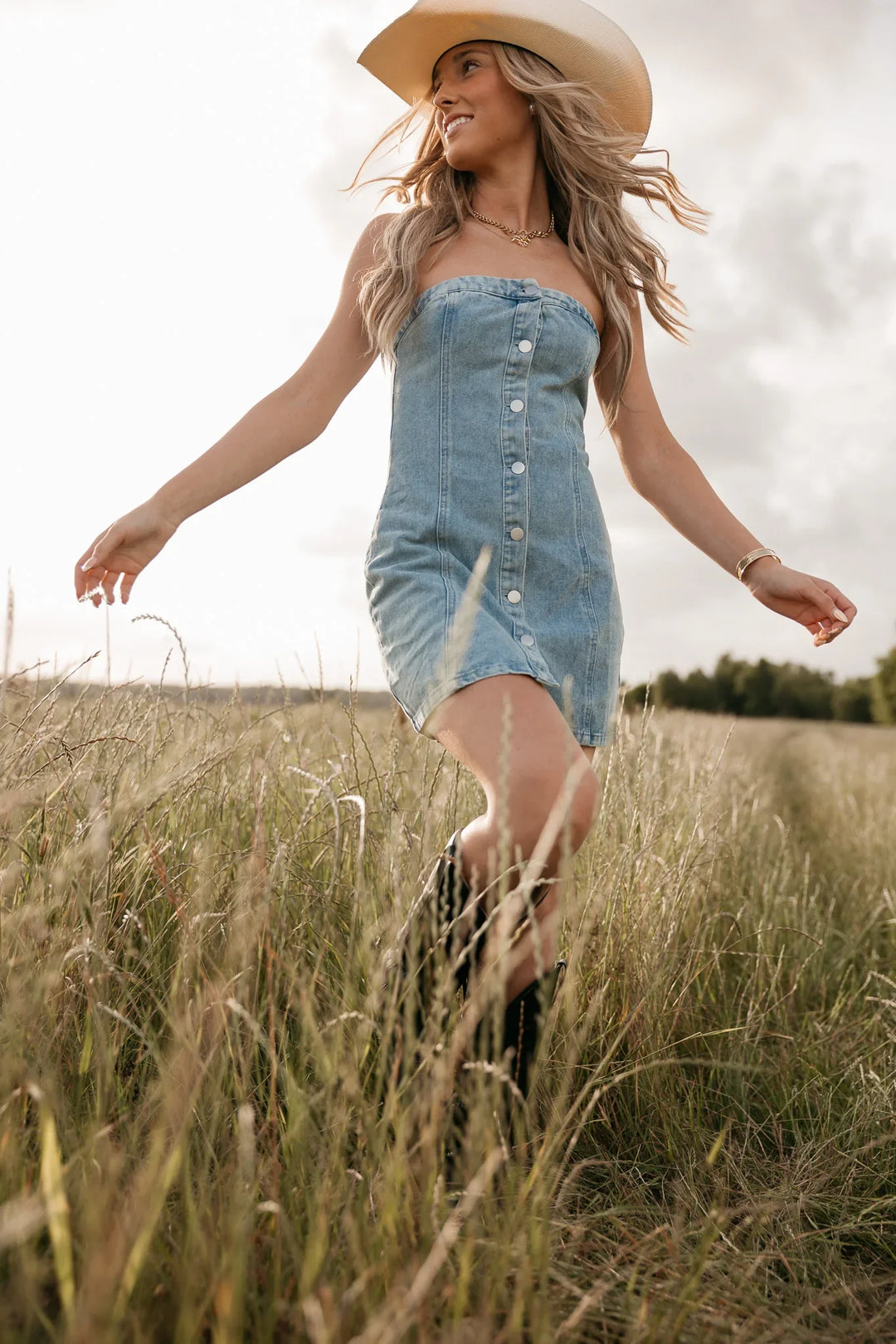 Woman in a denim dress and straw hat walking through a field.