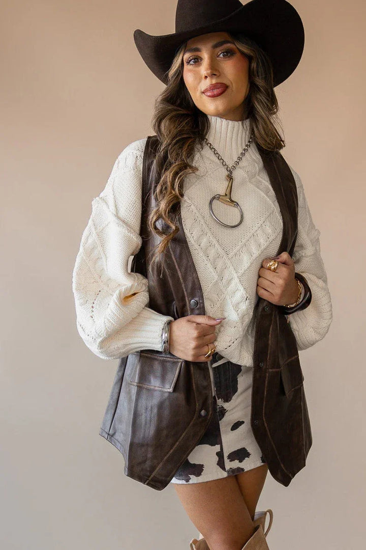 Woman in cowgirl hat, leather vest, cream sweater, and cow print skirt styled western boho