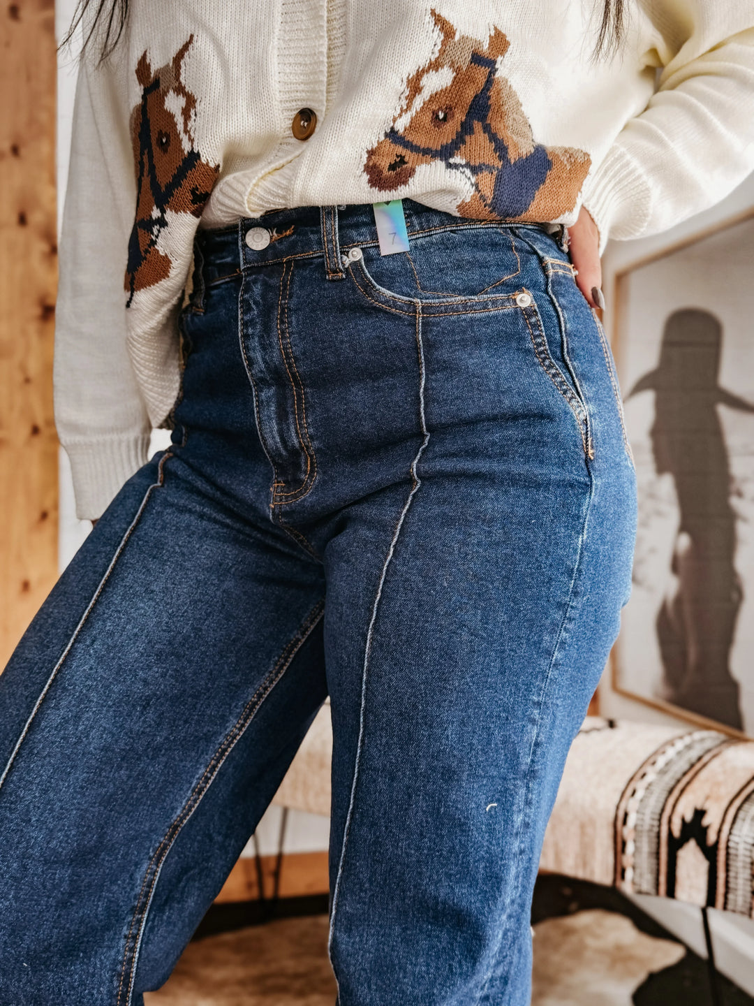 Woman in high-waisted western jeans and cream sweater with horse print, boho decor background