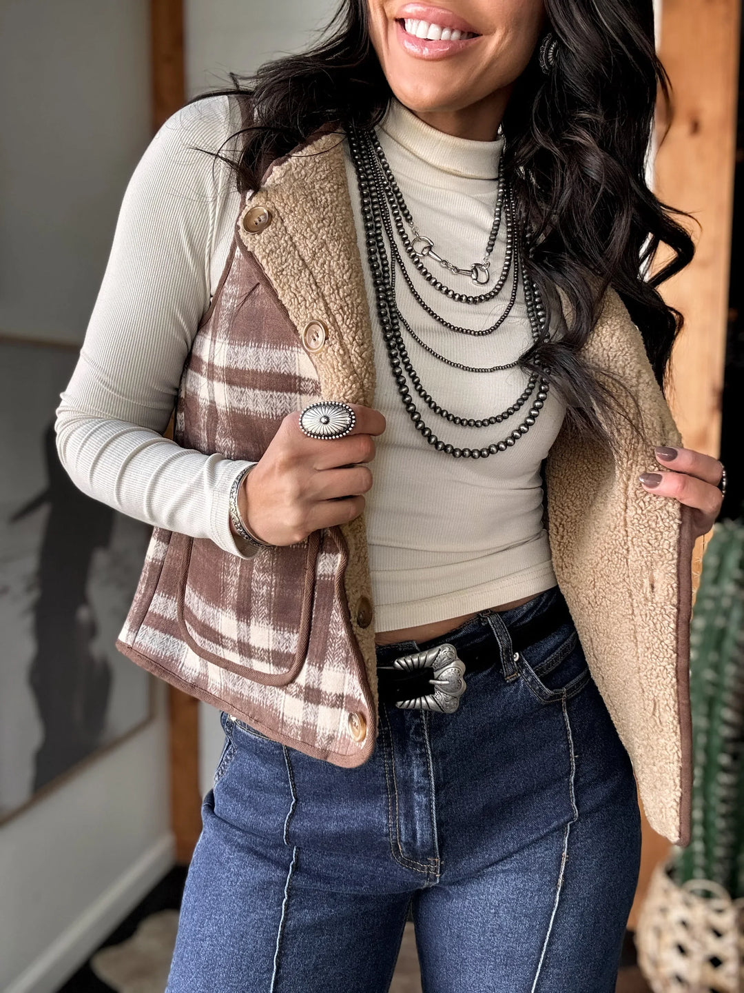 Woman wearing a plaid vest over a turtleneck with layered necklaces, smiling indoors.