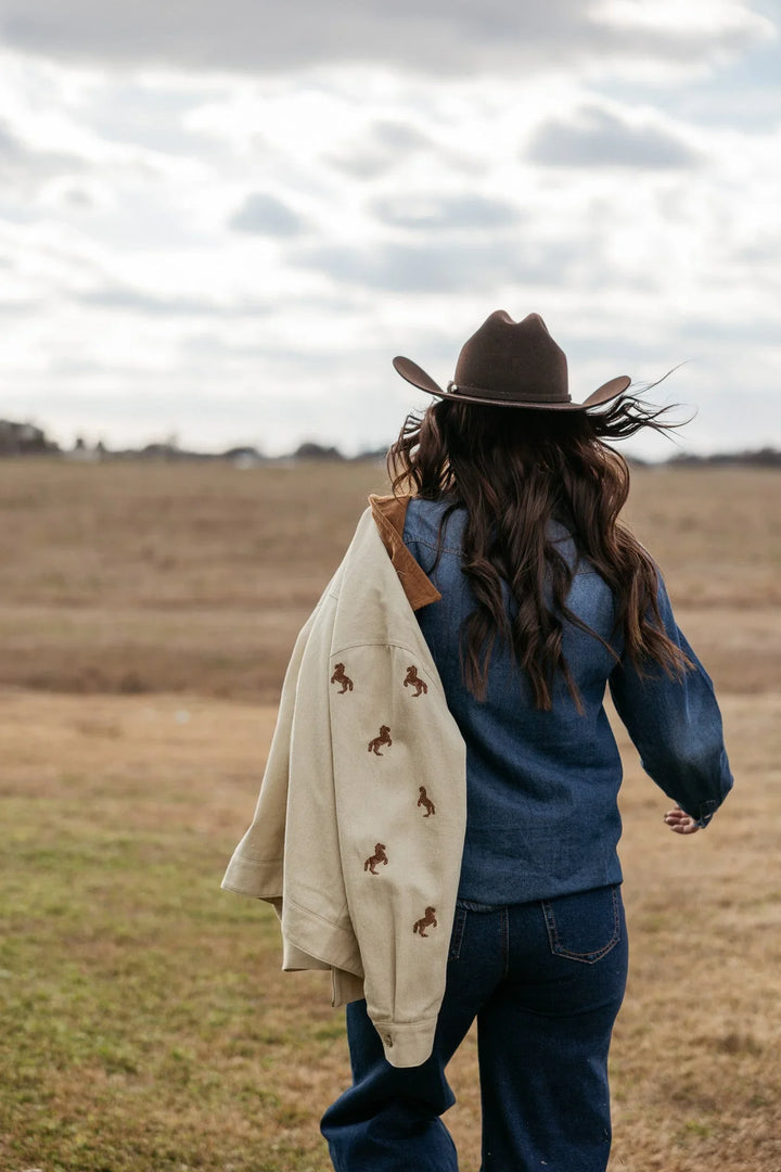 Woman in cowboy hat and denim outfit outdoors, holding tan jacket with horse embroidery, western style