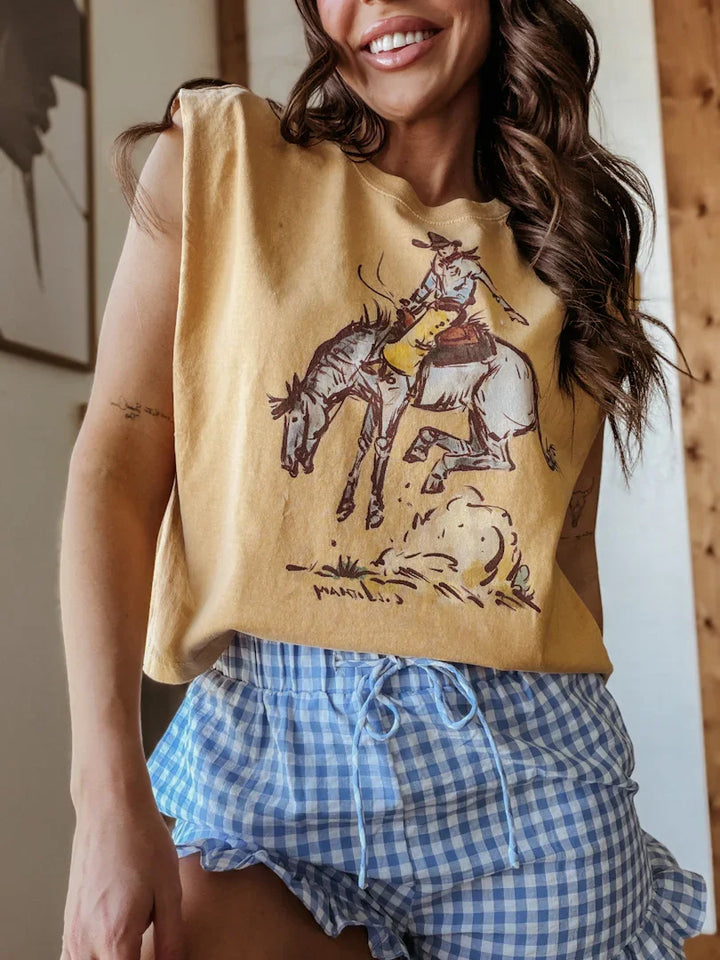 Woman wearing a mustard yellow sleeveless top with horse and rider design, paired with blue checkered shorts.
