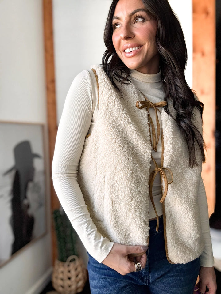 Woman wearing a cream sherpa vest with leather straps indoors.