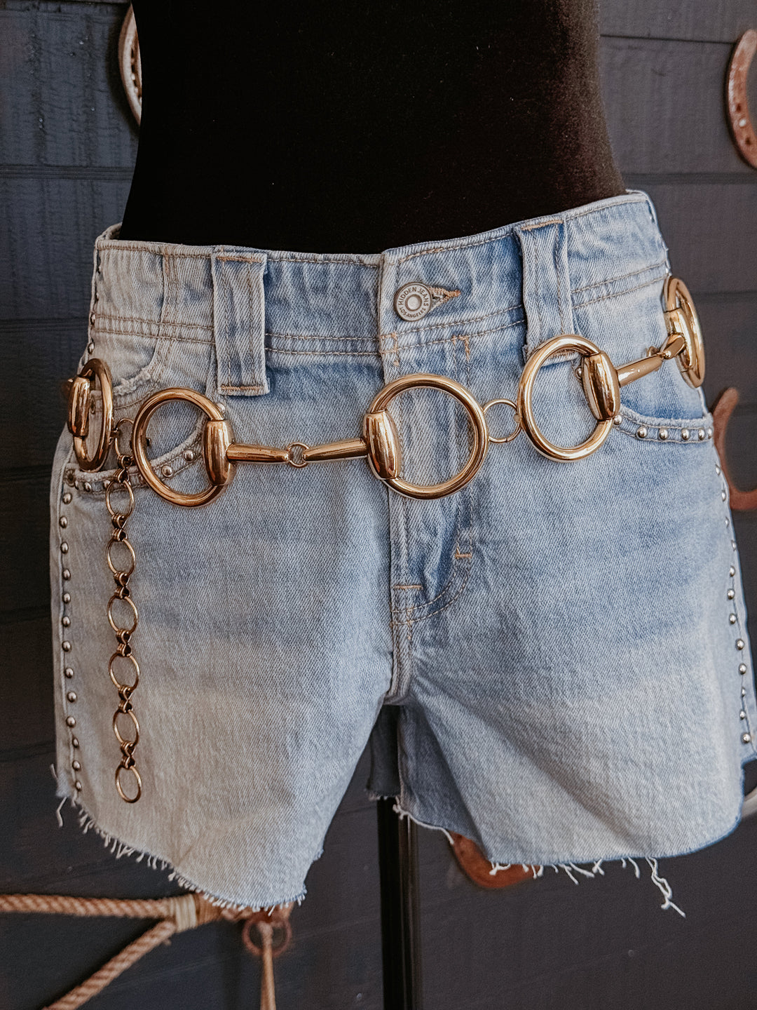 Denim shorts with a gold equestrian-style belt on a mannequin against a dark background