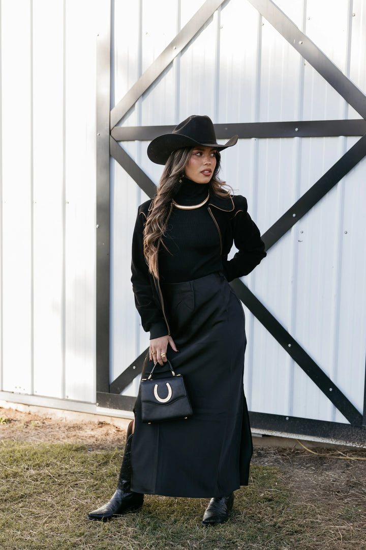 Woman in black cowgirl outfit with wide-brim hat, maxi skirt, and horseshoe purse standing by barn