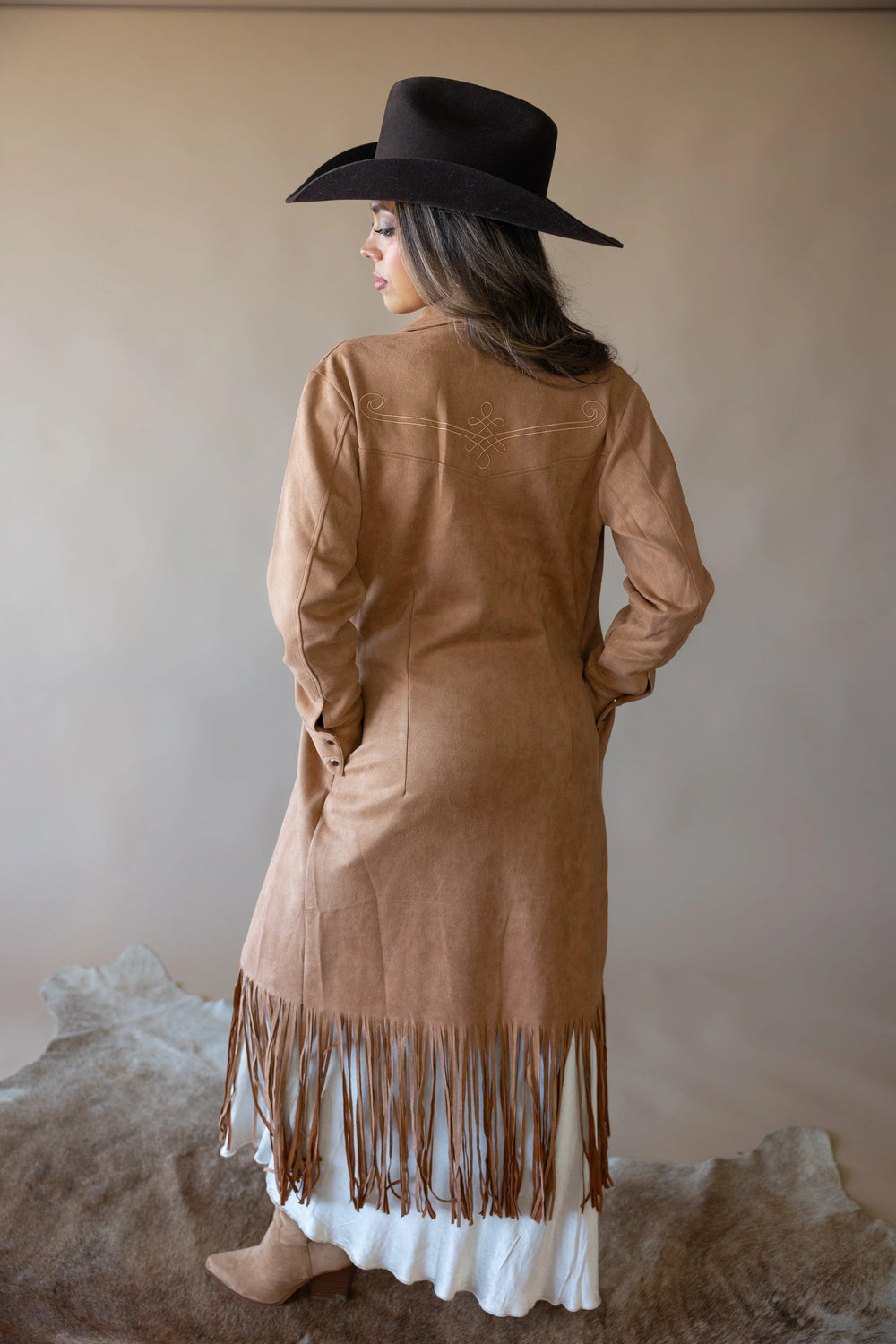 Woman in suede fringe western duster coat, black cowboy hat, and boots on cowhide rug