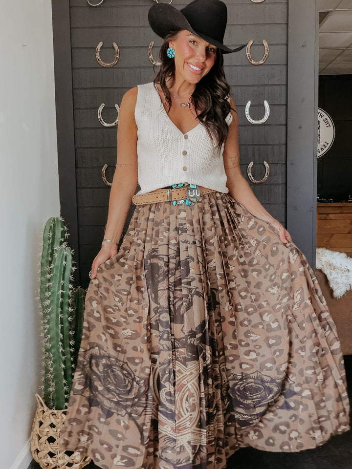 Woman wearing a white sleeveless top and patterned skirt with a black cowboy hat, standing in front of a wall with metal horseshoe decorations.