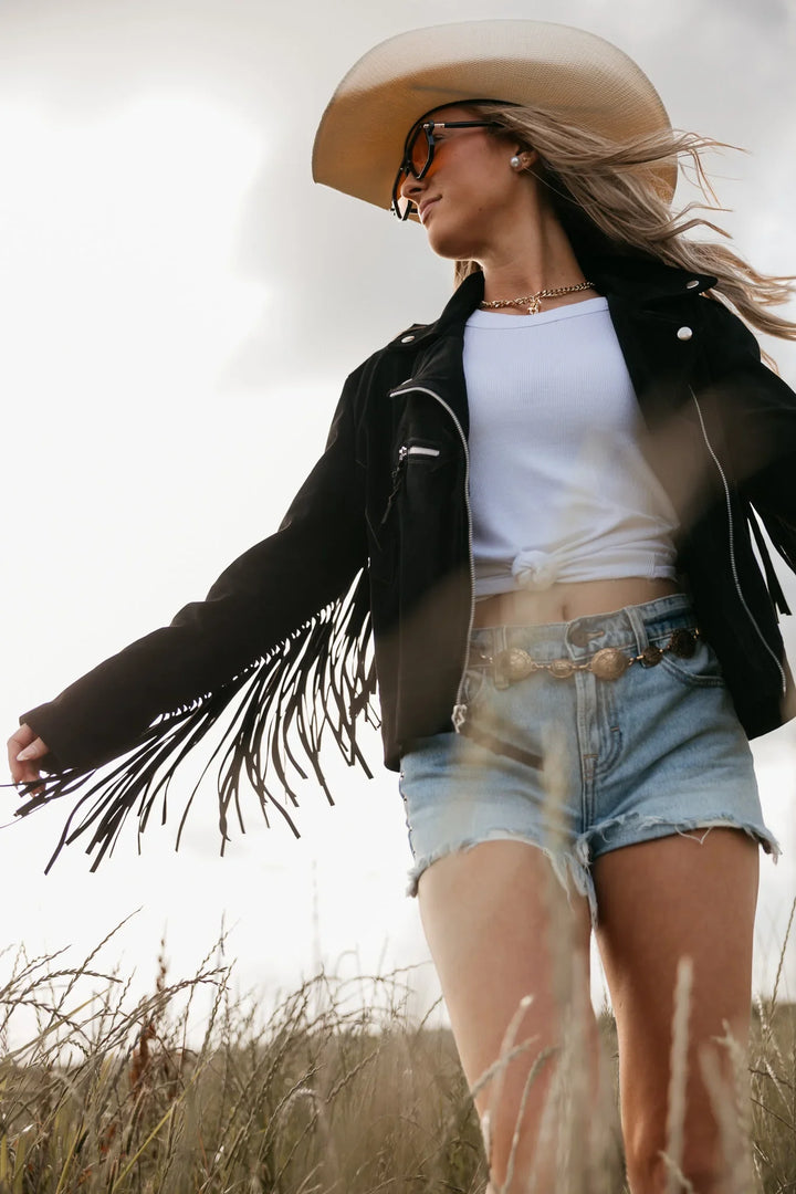 Person wearing a cowboy hat, black jacket with fringes, white top, and denim shorts in an outdoor setting.