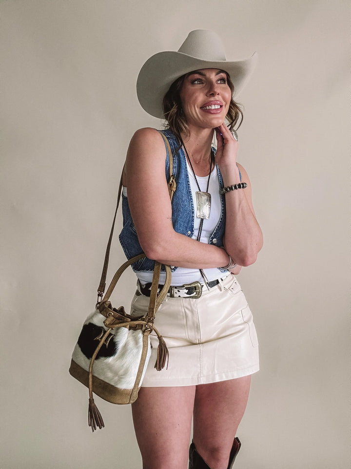 Smiling woman in cowboy hat, denim vest, cream mini skirt, Western bag and jewelry.