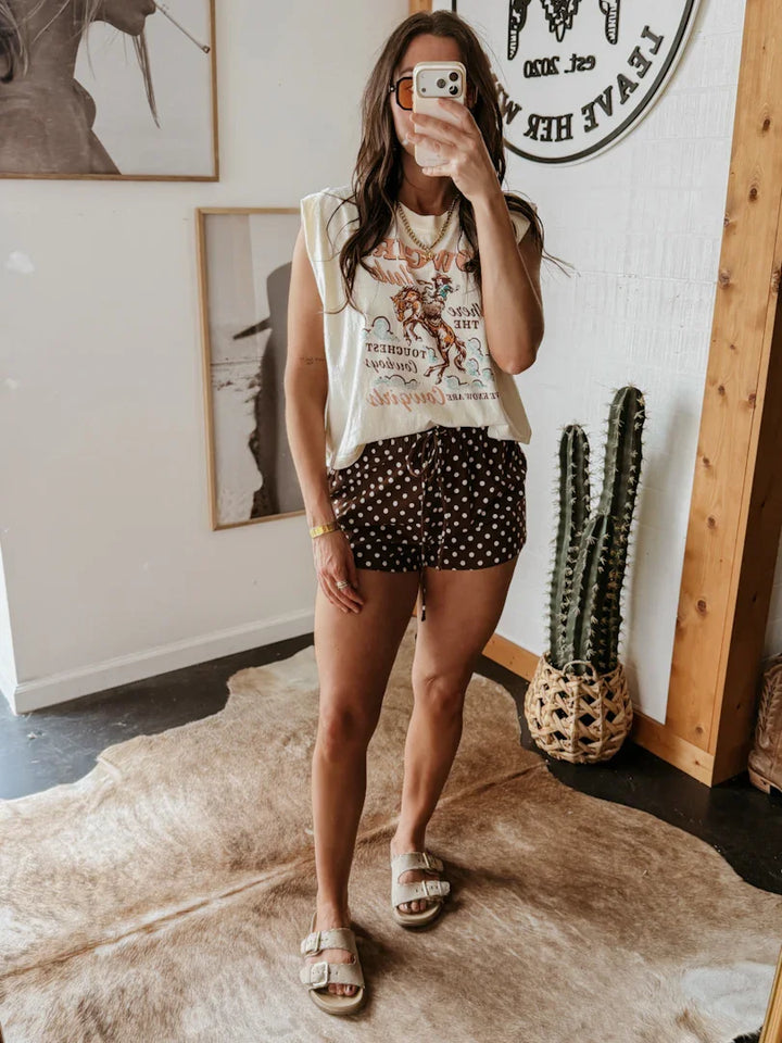 Woman taking a mirror selfie wearing a graphic t-shirt and polka dot shorts in a room with a cactus plant.