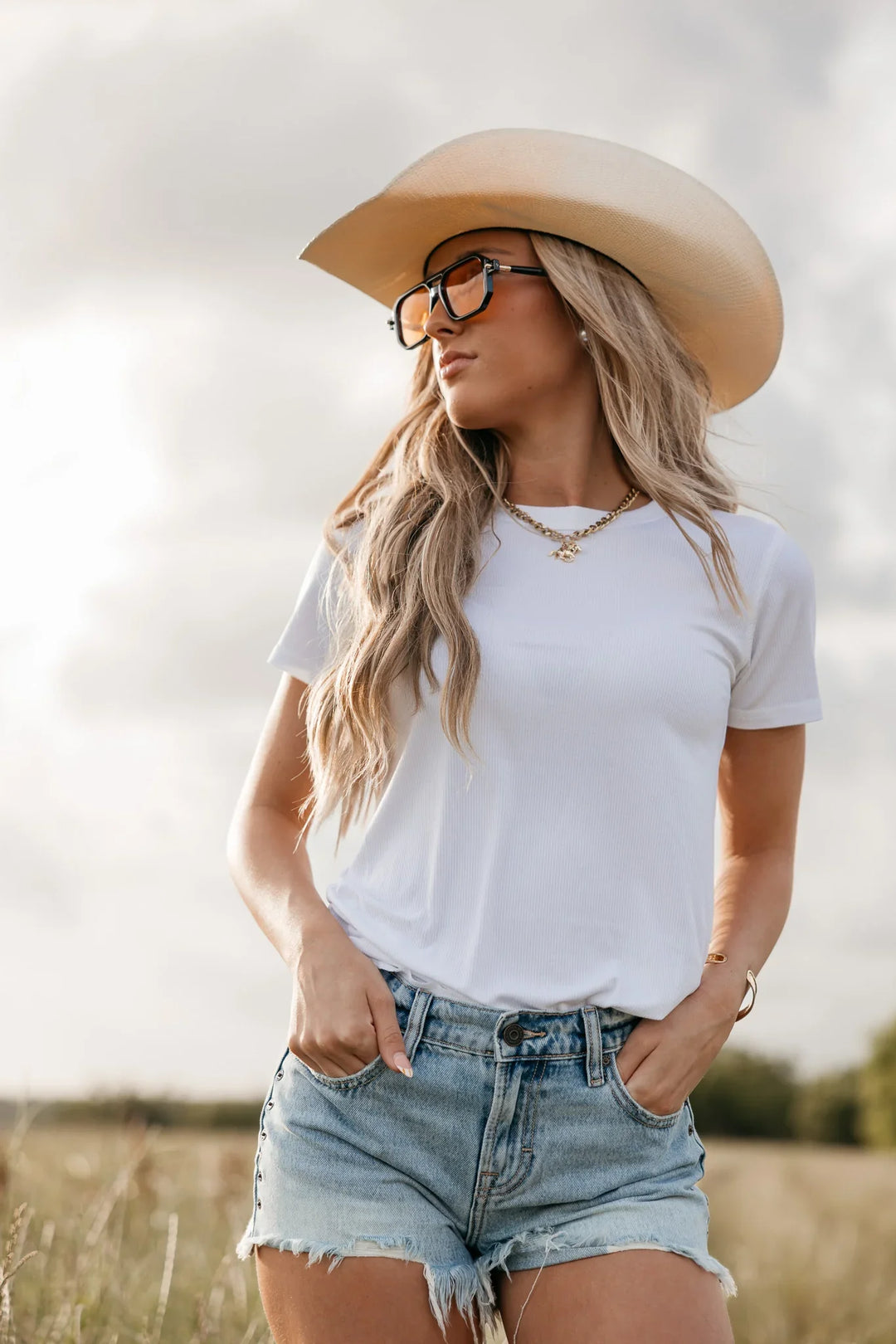 Woman wearing a white t-shirt, denim shorts, and a beige cowboy hat in a field.
