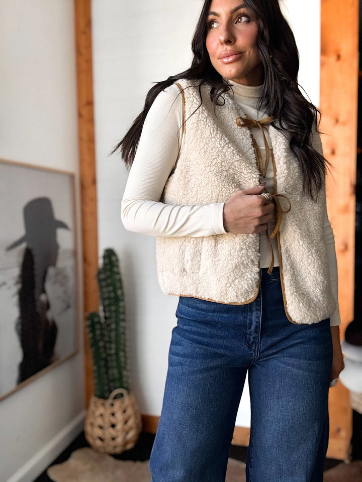 Woman wearing a cream vest over a white shirt and blue jeans in a room with a cactus and framed picture.