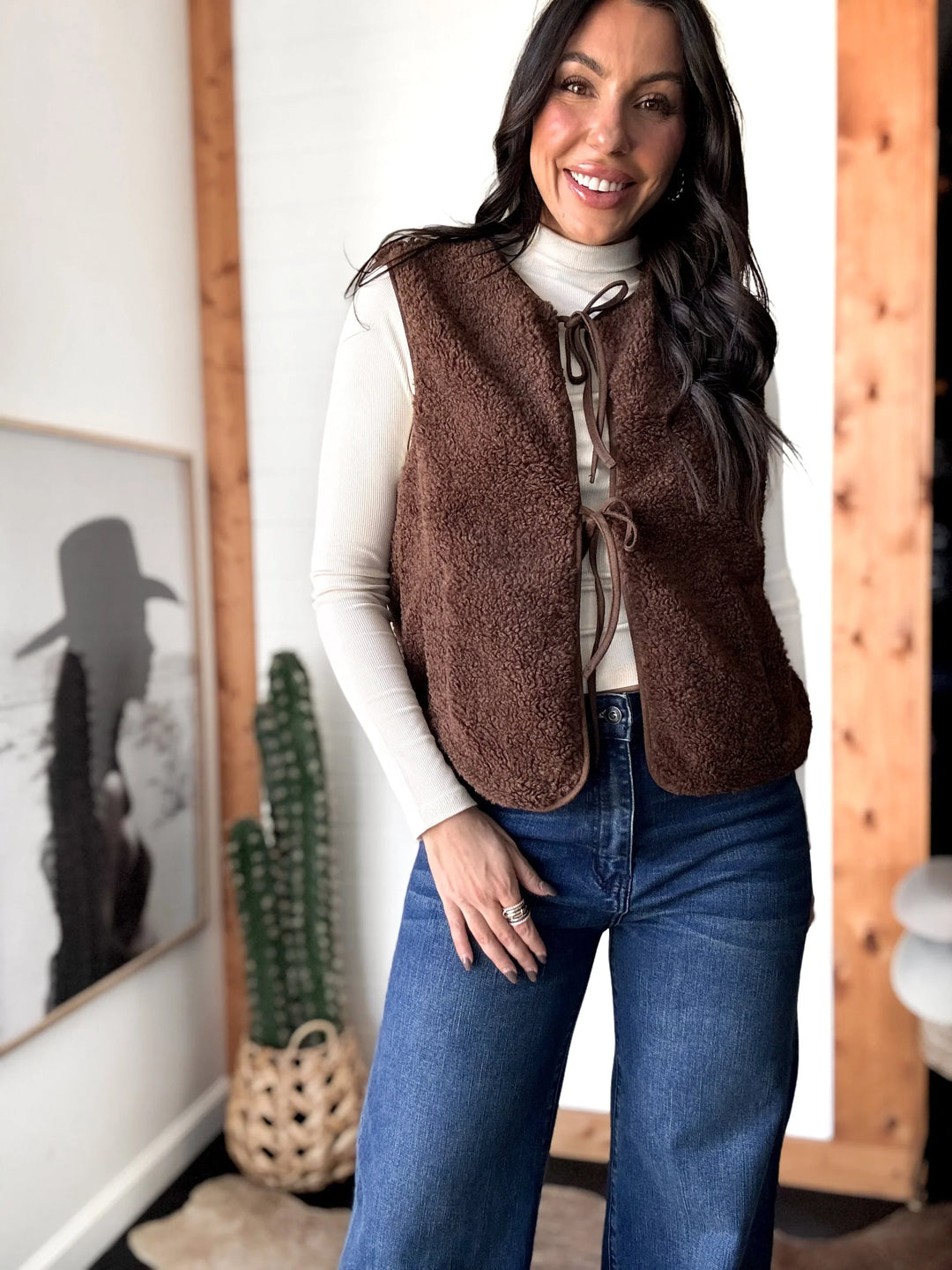 Woman wearing a brown vest over a white shirt and blue jeans indoors.