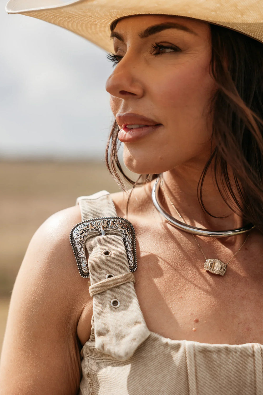Woman wearing a beige hat and overalls with a necklace, standing outdoors.