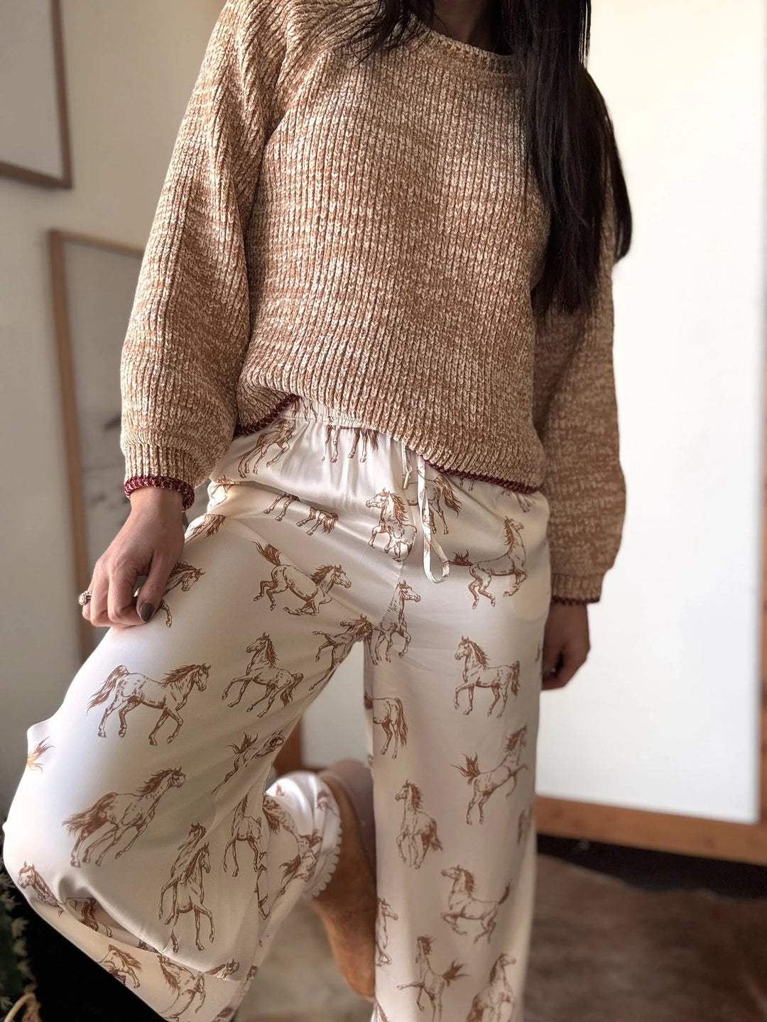 Person wearing a beige knit sweater and white pants with horse prints indoors.