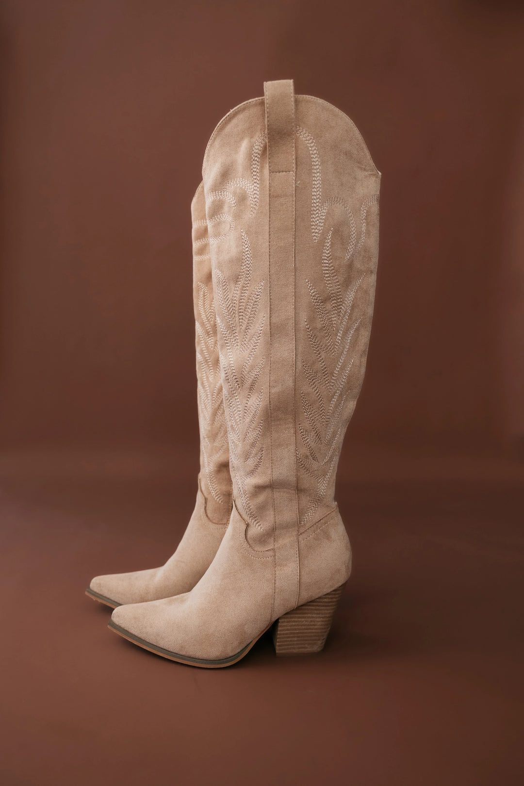 Tan suede western cowgirl boots with embroidery and block heels on brown background