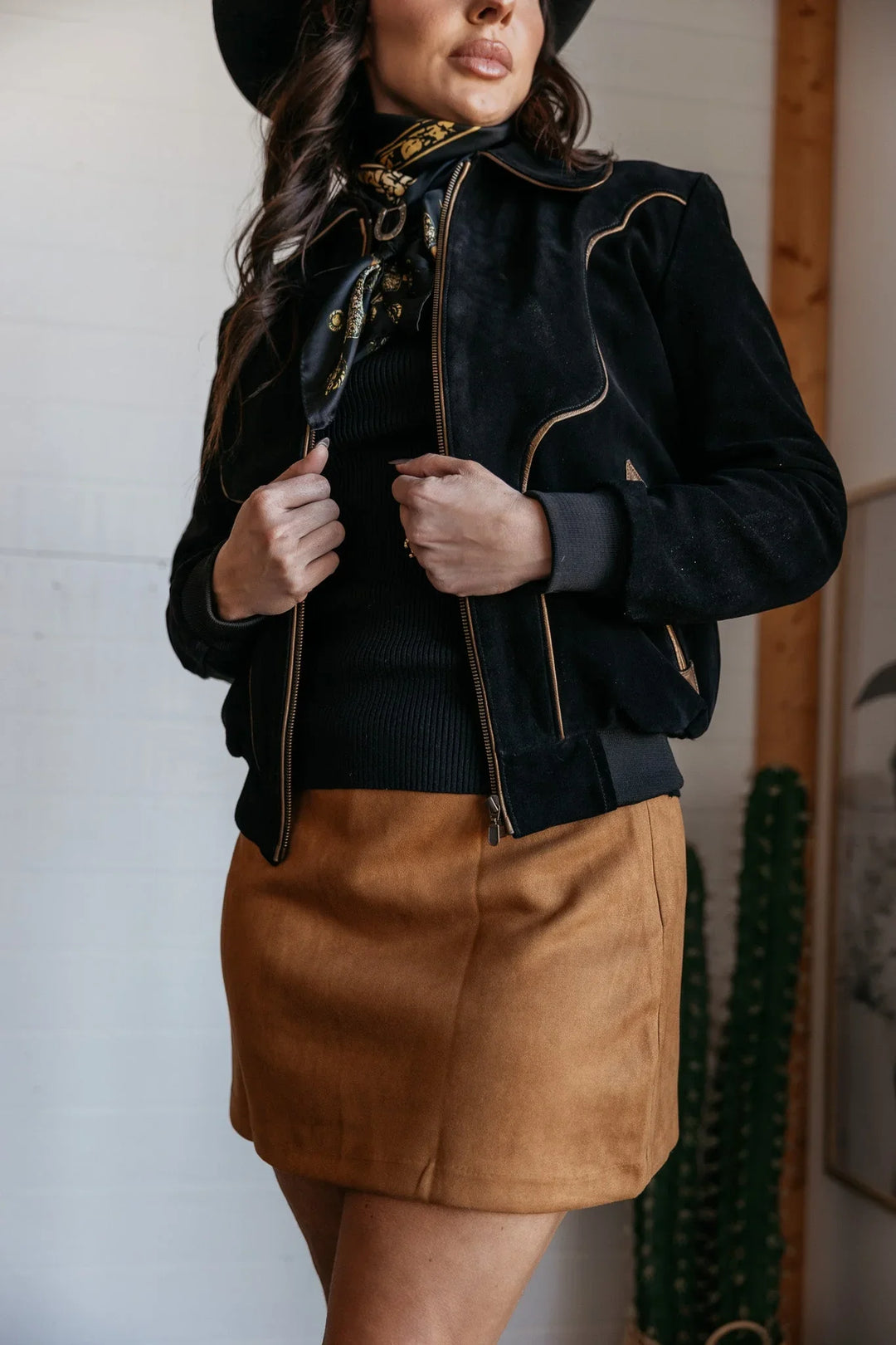 Woman in western boho outfit with black suede jacket, brown mini skirt, and black hat