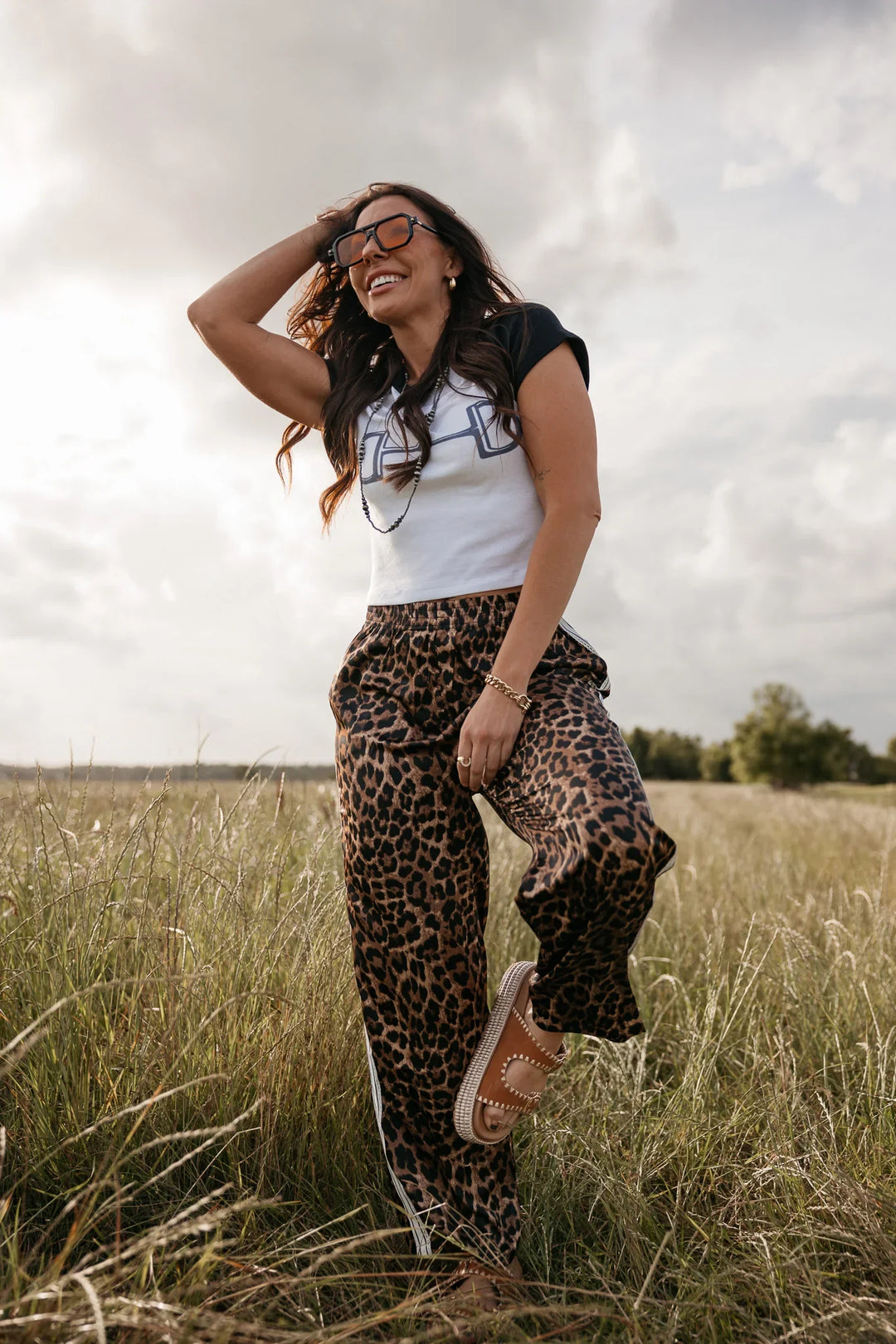 Woman in leopard print pants and white shirt standing in a field with a cloudy sky.