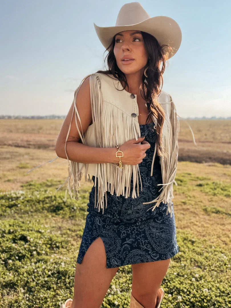 Woman in cowgirl hat, cream fringe vest, blue paisley dress and boots in a western field