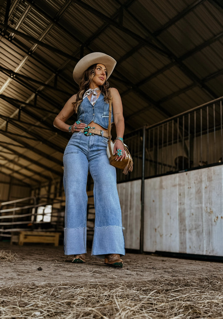 Woman in a western denim outfit with flared jeans, cowboy hat, and turquoise jewelry in a barn.