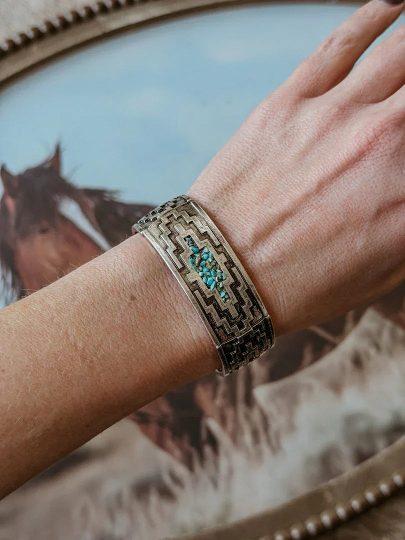 Hand wearing a decorative bracelet with turquoise stones against a background of horses.