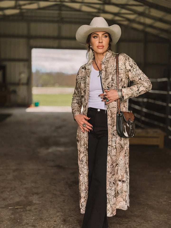 Woman in a western boho outfit with a cowboy hat, long patterned duster, and black flared pants standing in a barn.