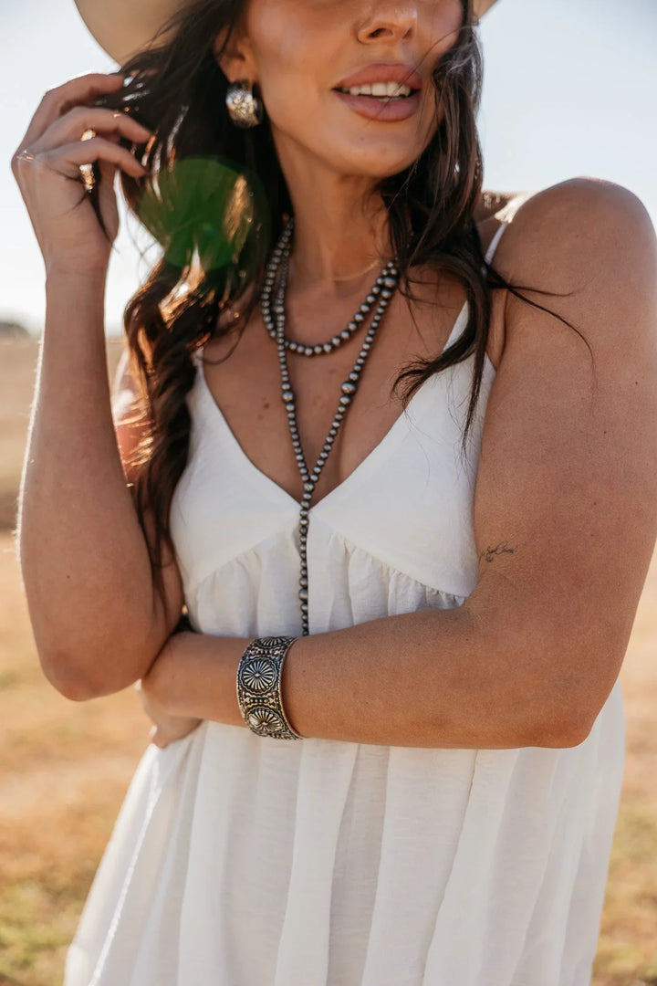 Woman in a white dress with layered necklaces and a watch, standing outdoors.