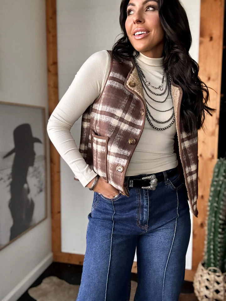 Woman wearing a plaid vest, white turtleneck, and blue jeans indoors.