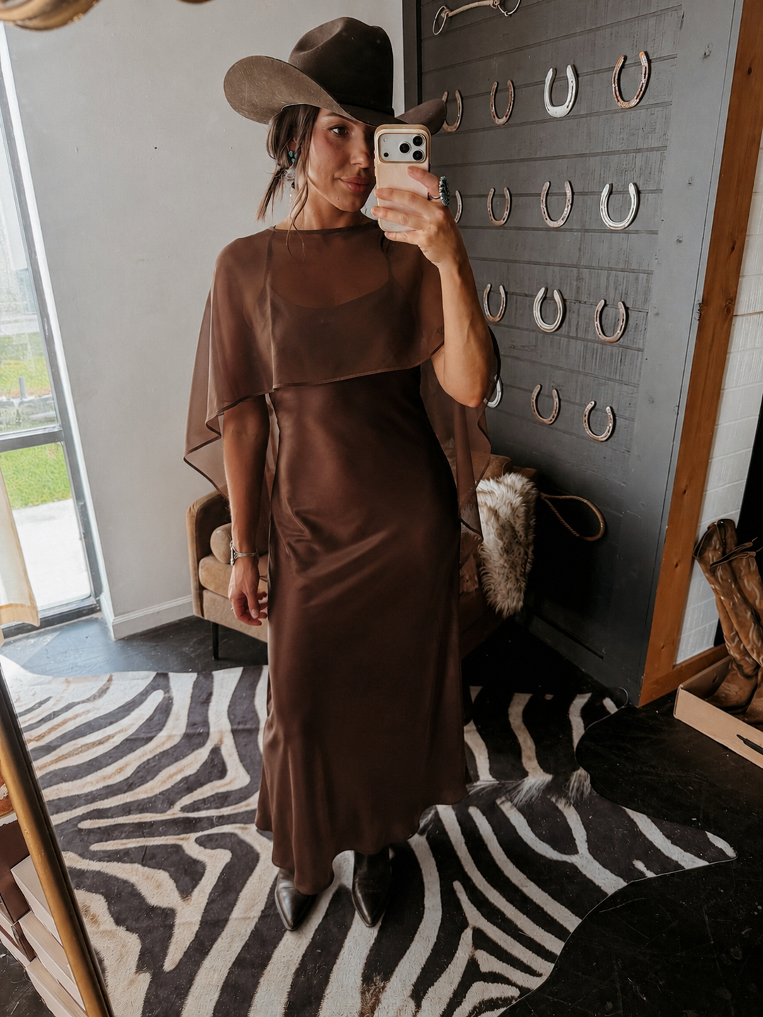 A woman takes a mirror selfie in a boutique wearing a brown midi dress with draped cape and a matching brown cowgirl hat.