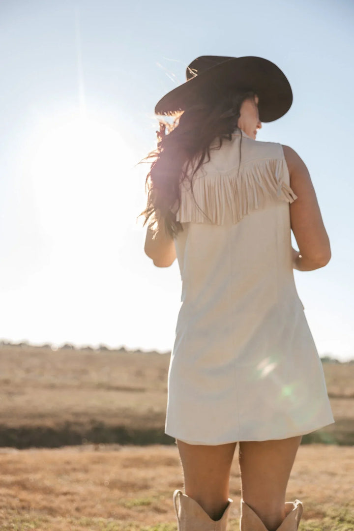 Woman in western fringe dress, cowboy hat, and boots standing outdoors in sunlight