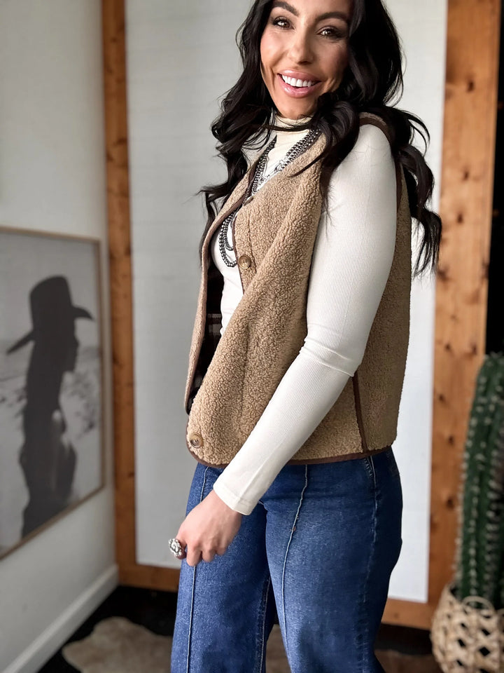 Woman wearing a brown vest over a white shirt and blue jeans indoors.