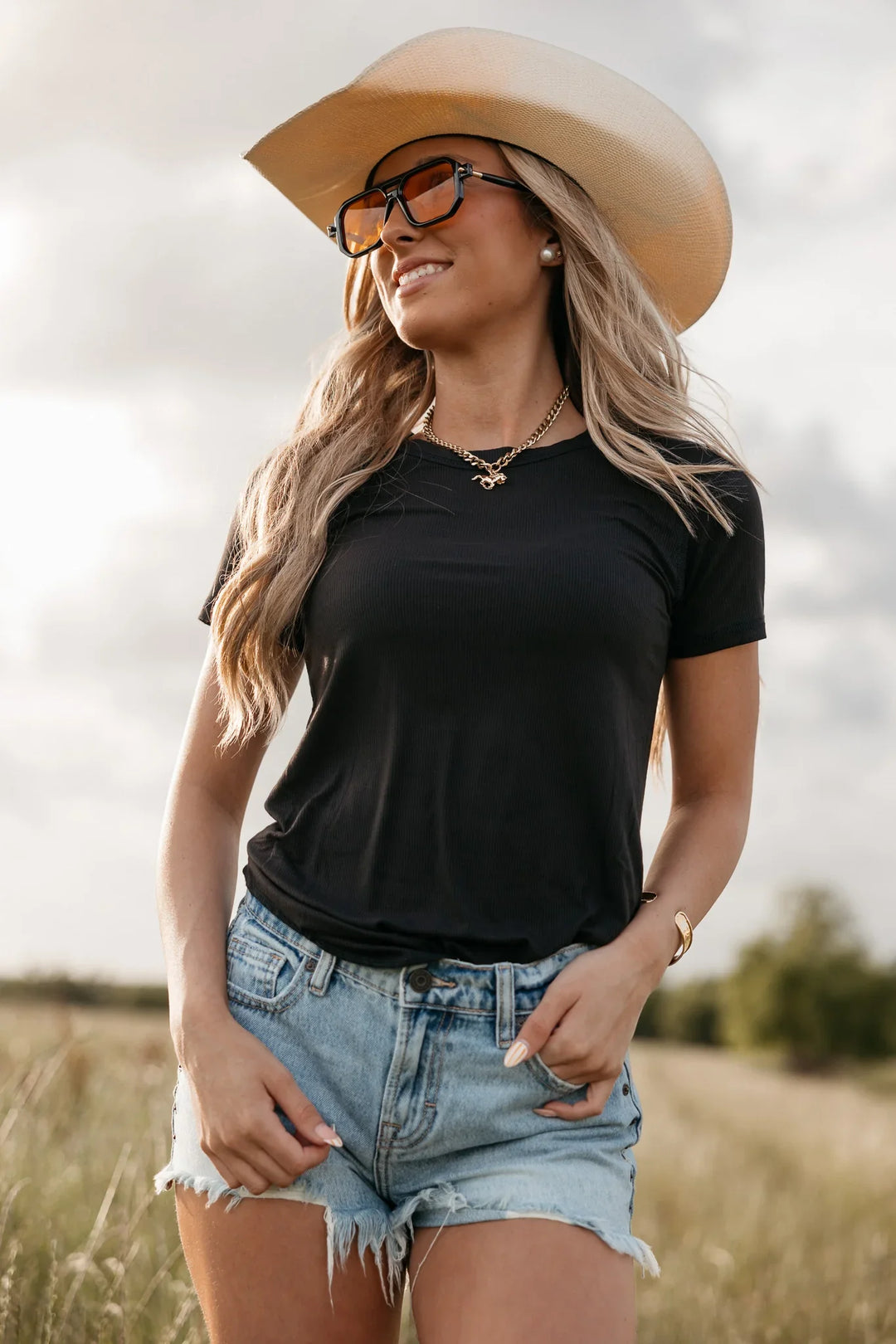 Woman wearing a black t-shirt, denim shorts, and a wide-brimmed hat in a field.