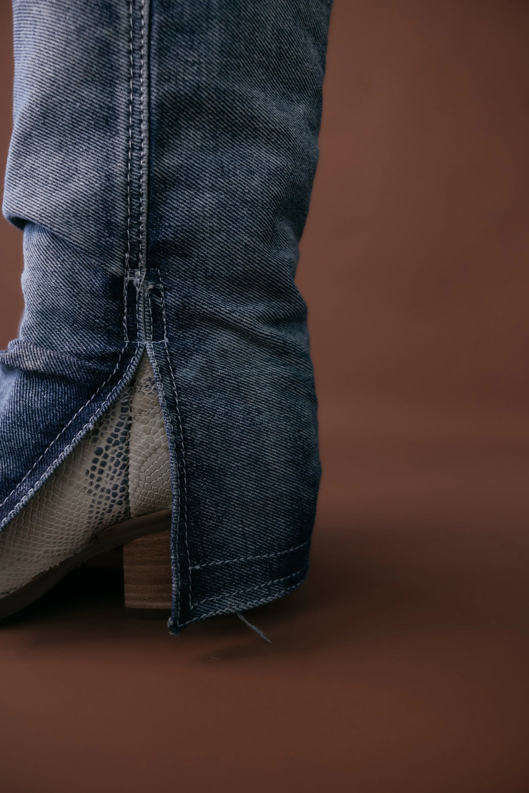 Close-up of a denim-covered western boot with snakeskin detail and wooden heel on brown background