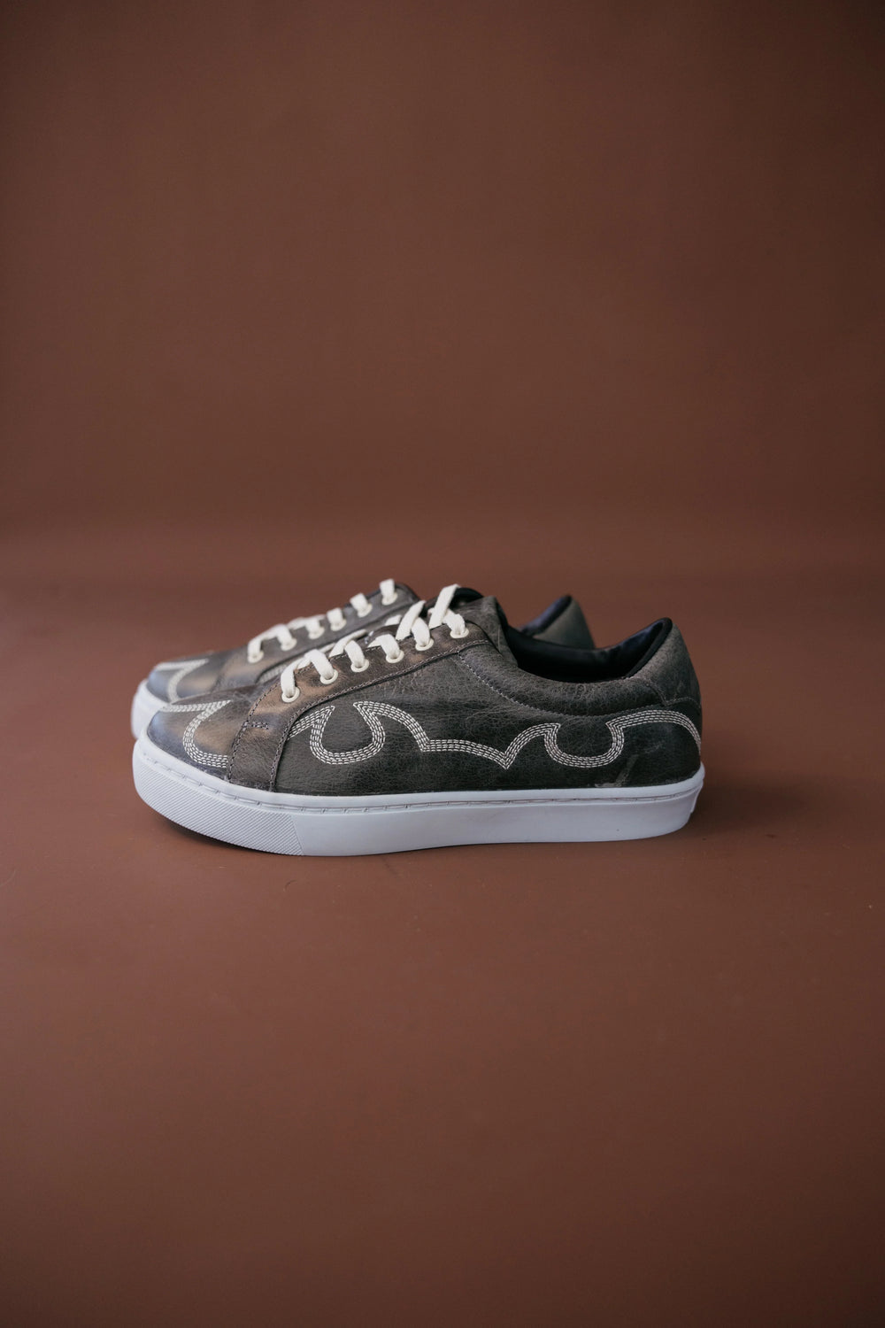 Western-inspired brown sneakers with decorative stitching and white soles on brown background