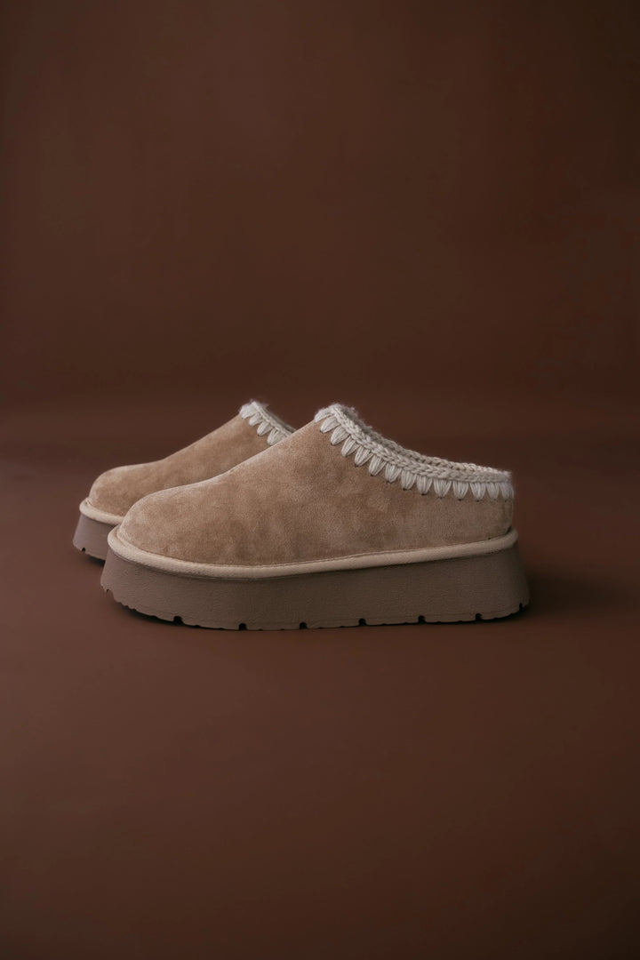 Pair of beige slippers with white trim on a brown background