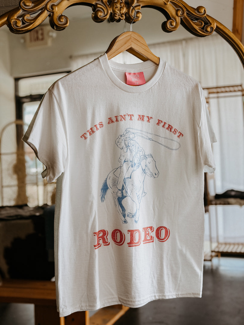 White t-shirt with red and blue graphic and text on a hanger in front of a mirror.