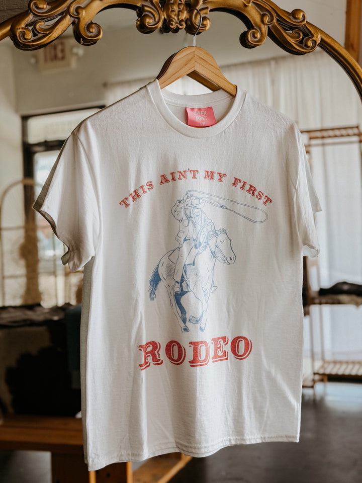 White t-shirt with red and blue graphic and text on a hanger in front of a mirror.