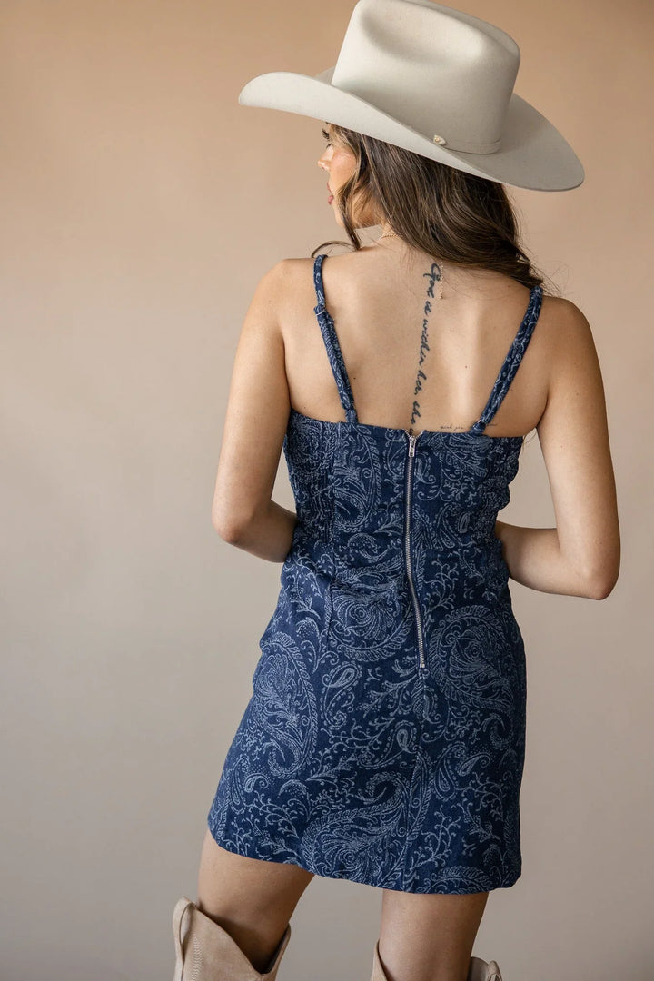 Woman in blue paisley mini dress, cream cowboy hat, and western boots, facing away
