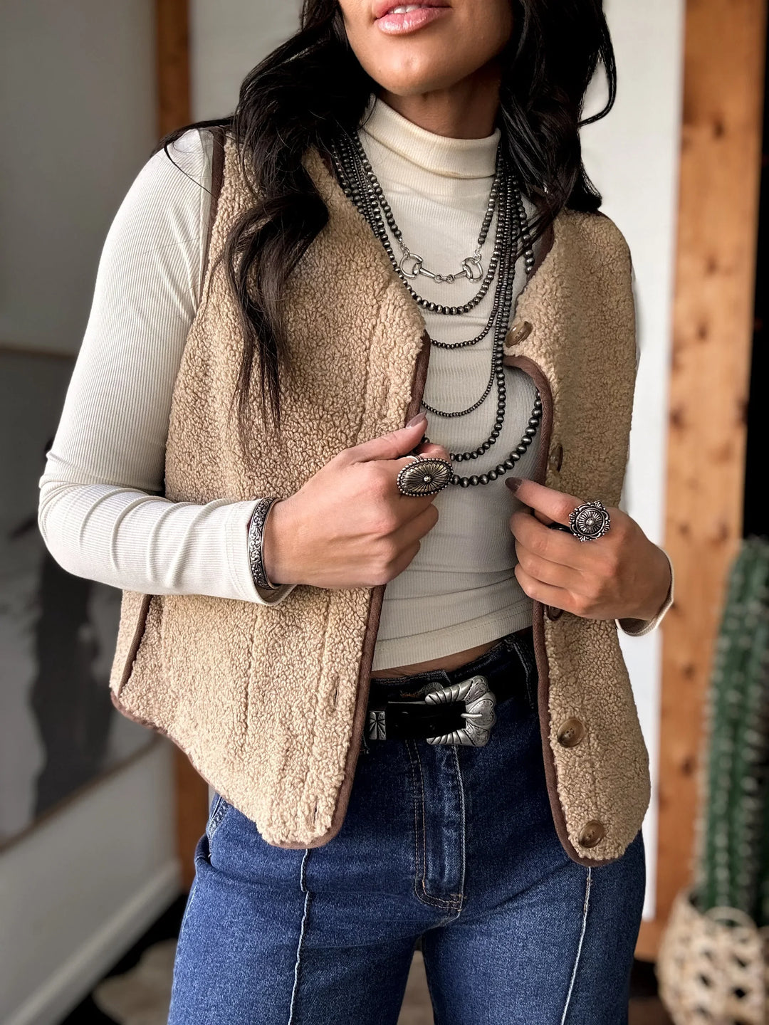 Woman wearing a beige vest over a white shirt with blue jeans indoors.