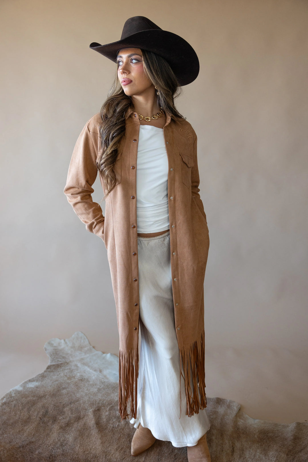 Woman in western boho outfit with tan fringe duster, cowboy hat, and white skirt