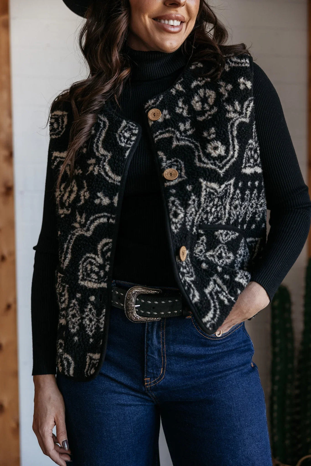 Woman wearing western boho patterned vest, black turtleneck, and blue jeans with belt