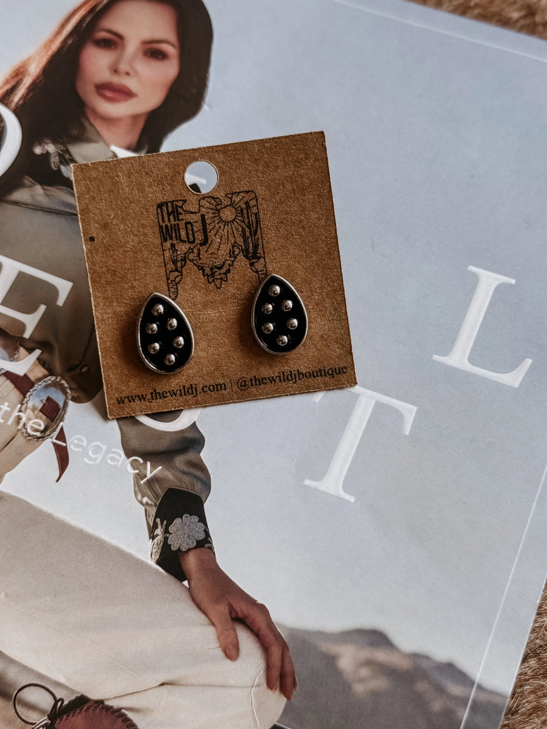 Black earrings on a brown card with 'the Wild' branding, displayed on a magazine background.