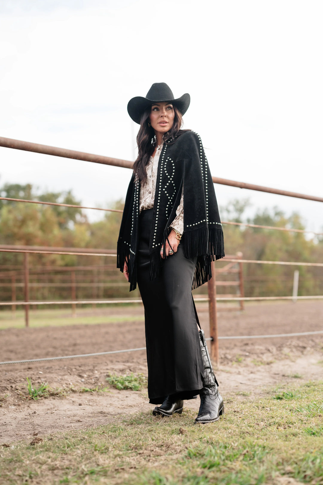 Woman in black cowgirl hat, fringe jacket, maxi skirt, and boots in a western ranch setting