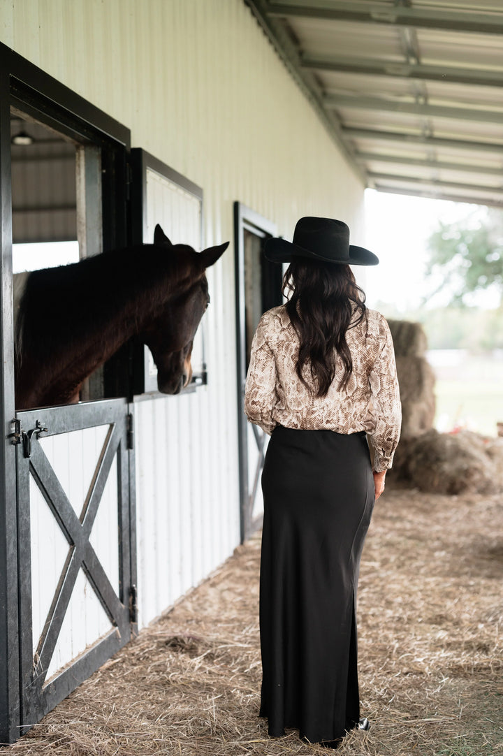 Woman in snakeskin blouse, black maxi skirt, and black hat standing by horse stable