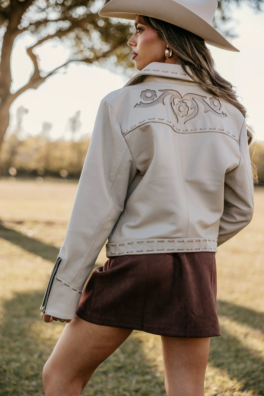 Woman in white western embroidered jacket, brown skirt, and cowgirl hat outdoors